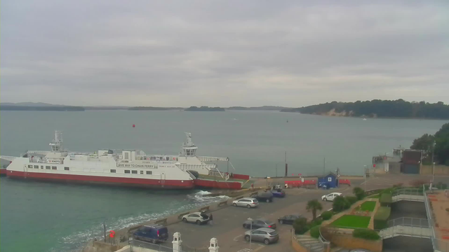 A view of a harbor with a large white and red ferry docked on the left. The water is calm, stretching out to distant islands under a cloudy sky. In the foreground, there is a parking area with several cars, and some greenery and pathways are visible. On the right, there are small buildings and a pier extending into the water.