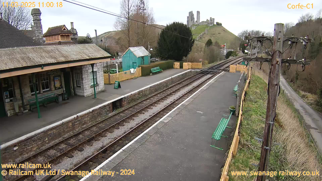 A view of a train station with a stone platform and wooden benches. In the background, there are green hills topped with ruins of a castle. To the left, there's a building with a sloped roof and a green door. Nearby, a small teal shed and a fence with a sign showing "WAY OUT." The railway tracks run alongside the platform, and a power pole is visible on the right. The sky is overcast.