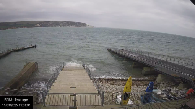 A coastal scene showing a rocky shoreline with concrete steps leading down to the water. To the left, a wooden pier extends into the sea, while on the right, another pier is visible. The water is choppy, with small waves, and the sky is overcast. Two kayaks, one yellow and one blue, are positioned next to the steps, alongside some equipment. The background features cliffs and greenery, partially obscured by clouds.