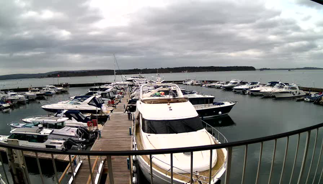 A view of a marina with numerous boats moored in a calm waterway. In the foreground, a large white yacht is docked, surrounded by smaller boats of various sizes and colors. The scene is set under a cloudy sky, with hints of land in the background. The dock is made of wooden planks, extending into the water where more boats are visible.