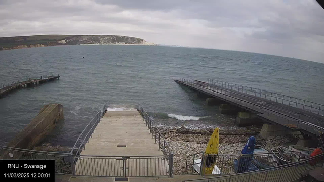 A coastal view showing a rocky shoreline with steps leading down to the water. To the left, there is a short pier extending into the sea. In the foreground, two kayaks are resting on the stones, one yellow and one blue. The sea is choppy with gentle waves. A cloudy sky looms overhead, and a cliff can be seen in the distance on the right side of the image. The overall mood is overcast and serene.