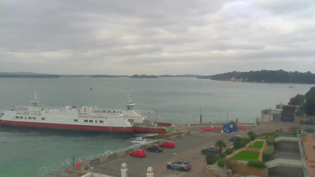 A cloudy waterfront scene featuring a large ferry with a white upper deck and red lower deck docked at a pier. Several red cars are parked in the foreground near the water. The sea is calm with gentle waves, and small boats can be seen in the distance. There are green trees and hilly land on the opposite shore.