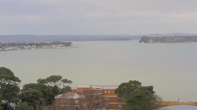 A panoramic view of a calm waterway bordered by an assortment of buildings and trees. In the foreground, there is a brown building with a flat roof, surrounded by greenery. The water stretches out into the distance, where sailboats and small vessels are visible. On the opposite shore, houses and a marina can be seen, with a backdrop of soft hills under a cloudy sky, indicating overcast weather.