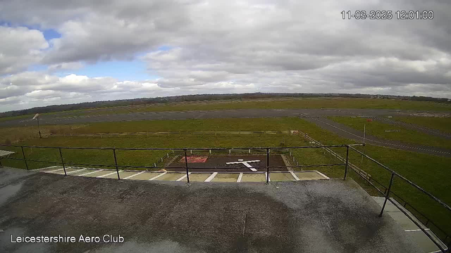 A view from a webcam of an open landscape with a large grassy area and a runway in the middle. There are gray clouds in the sky, and the scene appears to be overcast. In the foreground, there is a flat rooftop with a railing along the edge. A white cross shape is marked on the ground below, indicating a helipad area. To the right, a strip of dark pavement leads towards the horizon, where more green fields can be seen in the distance.