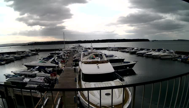 A marina filled with various boats is shown in the image. In the foreground, a large white yacht is docked next to a wooden walkway. Surrounding it are several smaller boats, some grouped together on the left and right sides. The water is calm, reflecting the overcast sky with gray clouds. In the background, there are land features and more boats on the horizon. The atmosphere appears tranquil, typical of a marina setting.