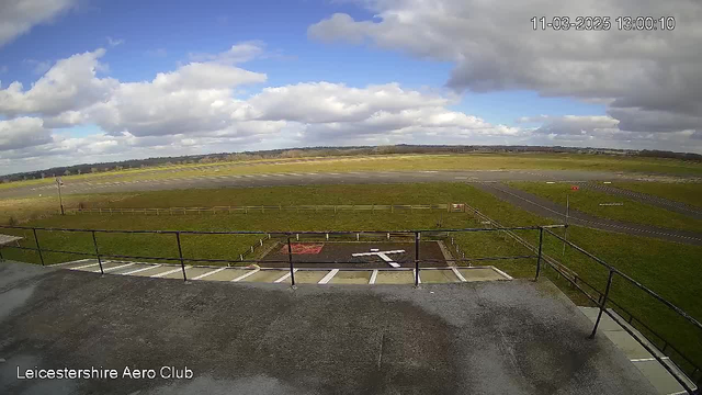 A view from a webcam showing a wide, open sky filled with clouds, floating above a grassy field that leads to a runway. The scene features a prominent white "X" marking on the ground, surrounded by a fenced area. The background displays a distant landscape with trees and a hint of a horizon. In the lower left corner, text indicates "Leicestershire Aero Club," and the date and time are displayed in the upper right corner.