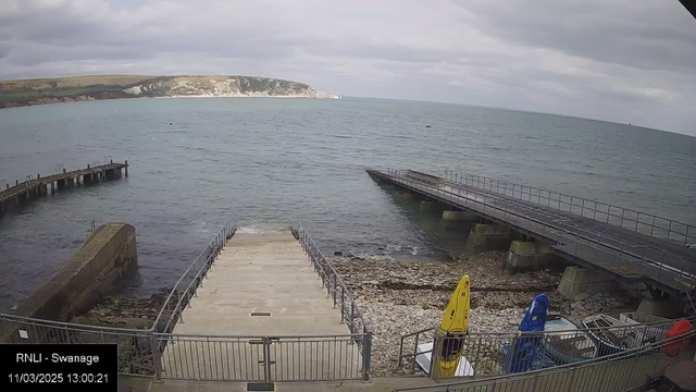 A rocky shoreline with a concrete ramp leading down to the water, flanked by a metal railing. To the right, there are two boats on the shore: one yellow kayak and one blue kayak. In the background, the calm sea stretches out towards a distant, grassy coastline with cliffs. The sky is overcast with gray clouds. A long wooden pier extends into the water from the left side of the image.