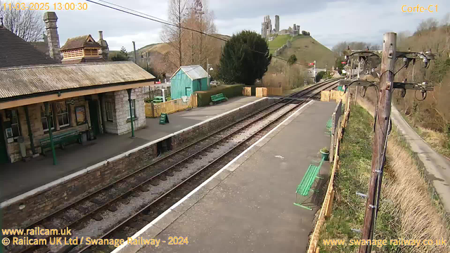 A railway station platform is visible, with green benches and a stone building with a slanted roof on the left. In the background, a hill rises with a castle-like structure atop it. The platform features two sets of railway tracks, and there are a few trees and a wooden fence surrounding the area. A green sign reads "Way Out." The sky is partly cloudy, creating a soft lighting effect over the scene.