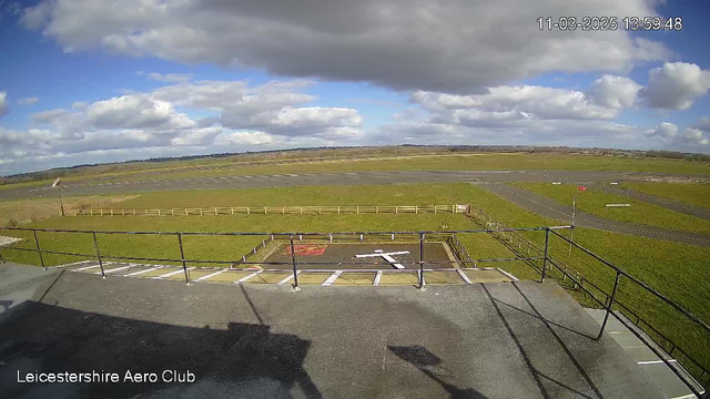 A panoramic view of an airfield under a partly cloudy sky. The foreground features a railing and a designated landing area marked with a white cross on the ground. In the background, there are patches of green grass and gray asphalt runways. The scene captures a wide expanse with low hills in the distance, indicating an open landscape. The time and date are displayed at the top of the image.