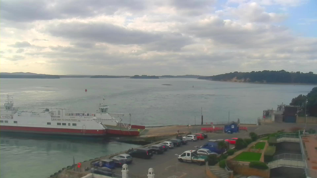 A view of a harbor with a ferry docked. The ferry is white with red accents, and there are several vehicles parked along the waterfront. The water is calm, reflecting the cloudy sky above, which has varying shades of gray with some blue peeking through. On the opposite shore, there are green hills and cliffs. In the foreground, there are landscaped areas with bushes and grass.