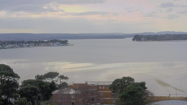 A view of a calm body of water reflecting soft pastel colors in the sky. In the distance, a coastline is visible with a row of buildings and boats. Closer to the foreground, there are trees on the left side and a brown building partially visible at the bottom. The landscape is serene and slightly overcast, with gentle hills in the background.