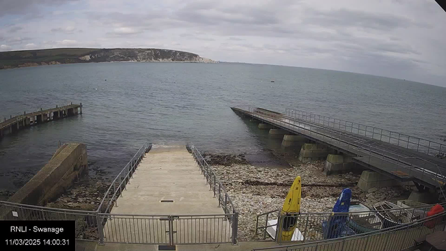 A view of a coastal area featuring a calm sea under a cloudy sky. In the foreground, there is a wide, concrete ramp leading down to the water, surrounded by a metal railing. To the left, a pier extends into the sea, with a section appearing slightly lower than the ramp. On the right, there are two colorful kayaks, one yellow and one blue, positioned near the edge of the ramp. The rocky shoreline is visible where the water meets the land. The distant land in the background includes cliffs and greenery.