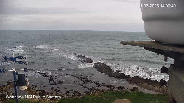 A coastal scene showing waves gently crashing on rocky shorelines. In the foreground, there is a weather vane with a blue arrow, indicating direction, along with a small bell-shaped structure. The ocean stretches into the distance, under a cloudy sky, with hints of grey and blue tones.