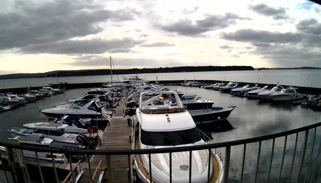 A marina filled with various boats and yachts is seen from a webcam. The scene features several boats docked closely together on calm waters. The sky is overcast with gray clouds, and the water reflects the subdued light. A wooden walkway leads along the docks, and there are distant hills visible on the horizon.