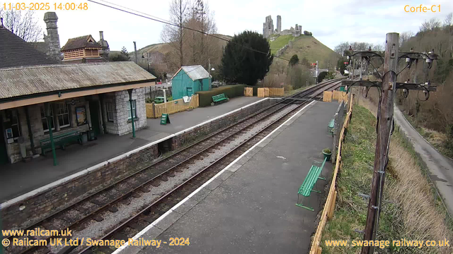 A quiet railway station scene featuring a stone platform with green benches. To the left, there is a rustic building with a sloped roof, and a blue-green wooden shed nearby. A sign reading "WAY OUT" is positioned on the platform. In the background, there are train tracks leading off into the distance. Above the platform, a tall pole with cables and a few small items can be seen. At the far end, a hill rises with ruins of a castle. The sky is mostly overcast, suggesting a calm, cloudy day.