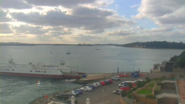 A scenic view of a harbor under a partly cloudy sky. In the foreground, a large white ferry with red accents is docked at the pier. Various cars are parked along the waterfront, including red and white vehicles. The calm water stretches out to the horizon, where a distant shoreline can be seen with greenery and cliffs. Small boats are also present on the water, and the overall atmosphere is tranquil.