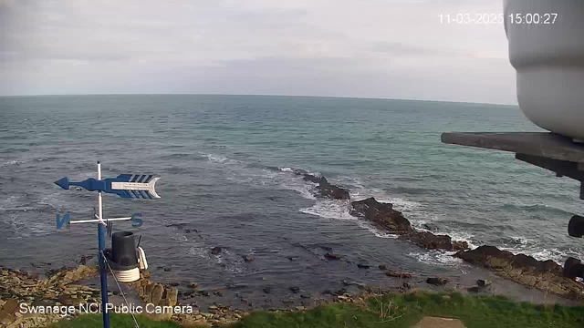 A coastal view captures the ocean meeting rocky shoreline under a cloudy sky. In the foreground, there's a weather vane pointing north, with a circular object partially visible. The water displays gentle waves, and some rocks are scattered along the coast. The scene has a serene and natural ambiance.