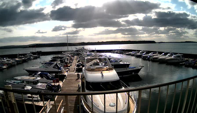 A scenic view of a marina filled with various boats and yachts moored on a calm water surface. The foreground features a large white motorboat with a shiny bow. The marina is lined with wooden docks leading to the boats. In the background, the water reflects a cloudy sky with sunlight peeking through, casting a shimmering light across the scene. The horizon is visible in the distance, with trees lining the shore. Overall, the atmosphere is tranquil and inviting.