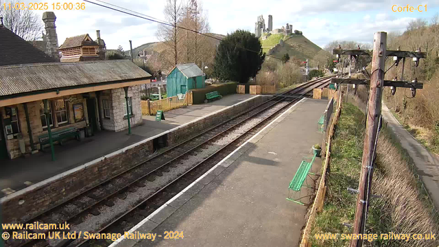 A railway station with a stone platform, featuring green benches and a small wooden shed. In the background, there is a view of a hill with a castle or ruins on top. The sky is partly cloudy, and the station has a rustic appearance with wooden fencing and trees nearby. Train tracks run parallel to the platform, and a sign indicating "WAY OUT" is visible.