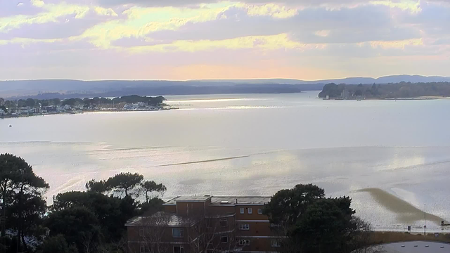 A wide view of a calm body of water extending to the horizon, reflecting soft light from the sky. In the foreground, there are some dark green trees and a brown building. The background shows a shoreline with several houses and boats, while distant hills are visible under a partly cloudy sky, painted in pastel colors.