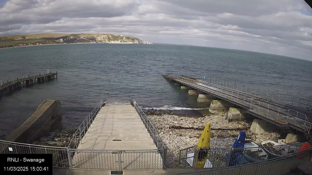 A view of a coastal area showing a partially visible marina and the ocean. There are two piers extending into the water; the one on the right is longer and has a railing. In the foreground, a concrete ramp leads to the water, surrounded by pebbles and small rocks. There are two kayaks, one yellow and one blue, nearby, along with several boats visible in the background. The sky is mostly cloudy with some patches of blue, and the cliffs can be seen in the distance along the coast.