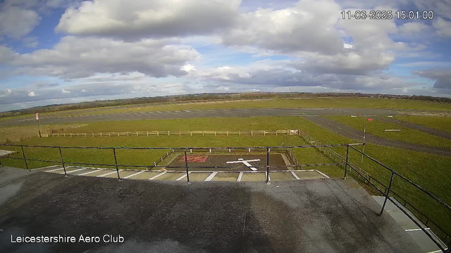 A panoramic view of an airport runway surrounded by green grass and a blue sky filled with fluffy clouds. In the foreground, there is a grassy area enclosed by a wooden fence, with a white markings of a cross on the ground, indicating a helicopter landing pad. The runway stretches out in the background, with a signpost visible on the left side, and various markings on the tarmac. The scene is bright and well-lit, showcasing a clear day at the Leicestershire Aero Club.