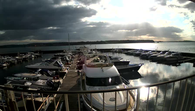 A marina scene filled with various boats docked in calm waters. In the foreground, a large white boat is tied to the dock. The background features additional boats of different sizes and designs. The sky is partly cloudy with patches of sunlight breaking through, reflecting off the water's surface. The overall atmosphere evokes a serene, peaceful harbor setting.