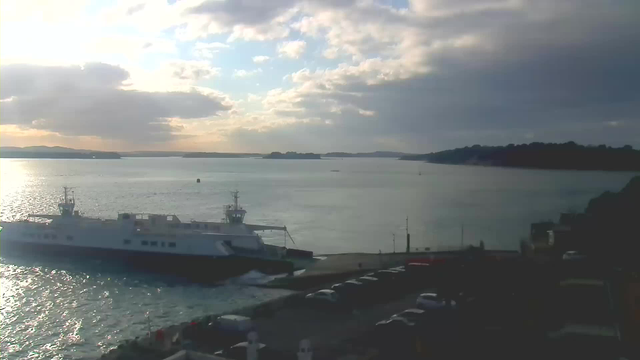 A ferry is docked at a harbor, with its bow pointed towards the camera. The water is calm, reflecting the soft light of the setting sun. In the background, hills and islands are visible, partially obscured by clouds. The sky shows a mix of blue and gray tones, indicating a change in weather. Several cars are parked along the quay, and the shoreline is lined with rocks.