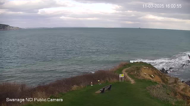 A view of a coastal area featuring a grassy bank that slopes down to the sea. The water is calm with gentle waves, reflecting the cloudy sky above. In the foreground, there's a bench placed on the grass facing the ocean. A warning sign is visible nearby, and the landscape is surrounded by sparse vegetation and rocky outcrops along the shoreline. The date and time are displayed in the top right corner.