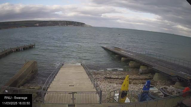 A view of a coastal area with dark water and a cloudy sky. In the foreground, there are two piers extending into the water; one pier is wooden and angled, while the other is concrete and flat. In front of the piers, there are steps leading down to the shore, which is lined with small stones and pebbles. To the right, there are two kayaks, one yellow and one blue, along with some equipment nearby. The background features a rocky shoreline and hills.