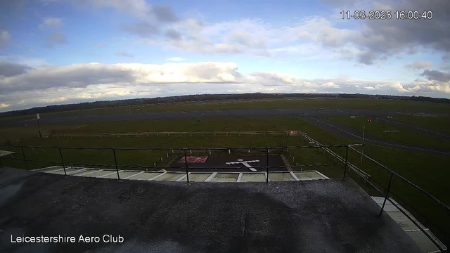 A view from a webcam at the Leicestershire Aero Club, capturing a landscape with a runway and grassy areas. The scene features a cloudy sky with varying shades of white and blue. In the foreground, there's a flat rooftop with a railing, and a marked area resembling a cross in white paint on a dark surface. The background shows a broad open area with a fence along the runway, which is mostly clear of aircraft.