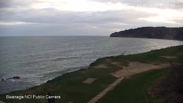 A coastal view showing the sea meeting the horizon under a cloudy sky. In the foreground, there is grassy land with a pathway made of stones leading towards a rocky coastline. Waves can be seen gently rolling in toward the shore, and the landscape appears quiet and serene.