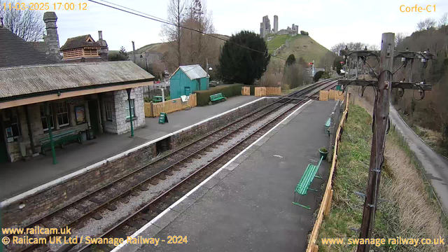 A railway station scene showing two parallel tracks leading into the distance. On the left, there is a stone building with a peaked roof, featuring large windows and a covered area with green benches. To the right, a wooden fence encloses a waiting area with additional green benches. A green sign reads "WAY OUT." In the background, a hill rises with remnants of a castle at the top, surrounded by trees and bare bushes. The sky is overcast, with a few clouds. A utility pole stands to the right, with various cables attached.