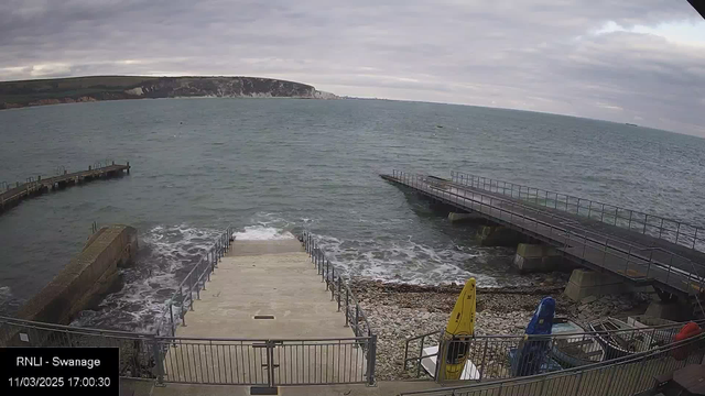 A coastal scene showing a stone staircase leading down to the water, with gentle waves lapping at the shore. To the right, a wooden pier extends into the sea, while on the left, a rocky jetty can be seen. In the foreground, there are several small boats stacked in a colorful arrangement, including yellow and blue kayaks. The sky is overcast with clouds, and the distant shoreline can be seen in the background.