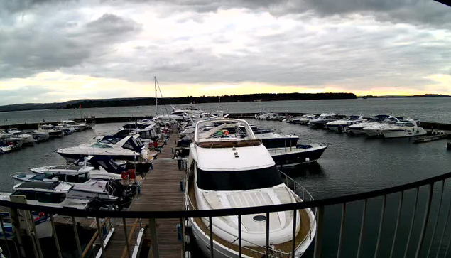 A marina filled with numerous boats docked in calm water. The foreground features a large white motorboat with a tan stripe. Surrounding boats are a mix of sizes and colors, predominantly white, blue, and black. There are wooden docks leading to the boats, with a railing in the foreground. The sky is overcast with gray clouds, and the horizon shows a hint of sunset with soft colors.