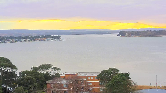 A serene view of a body of water at sunset, with a soft gradient of orange and yellow in the sky. In the foreground, there is a building with multiple stories, featuring a reddish-brown facade. To the left, a marina with boats is visible, alongside trees lining the water's edge. In the distance, hills are faintly visible under the colorful sky, creating a peaceful, natural scenery.