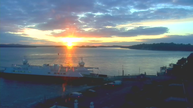 A scenic view of a body of water during sunrise, with the sun rising on the horizon, casting a warm golden glow across the sky and reflecting on the water. In the foreground, there are two boats docked near the shore; one larger ferry-like boat is prominently visible. The background features a line of hills partially covered by clouds under a sky with a mixture of blue and orange hues, indicating early morning light.