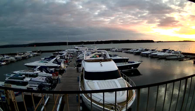 A marina filled with various boats and yachts is visible in the foreground. Some of the boats are moored closely together at wooden docks. A large white yacht is prominently positioned in the center, with a flat deck and a canopy. The water is calm, reflecting the colors of a sunset, where the sky displays shades of orange and gray. In the background, there are trees and hills silhouetted against the horizon. The scene has a tranquil ambiance, with scattered clouds adding texture to the sky.