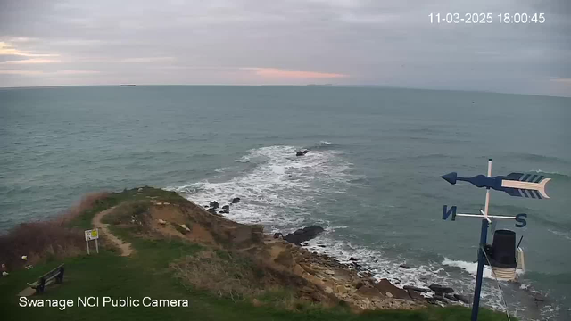 A coastal scene featuring rolling waves crashing against a rocky shore. In the foreground, there is a grassy area with a bench and a sign. To the right, a weather vane is visible, indicating directions. The sky is overcast with hints of color near the horizon, suggesting sunset. The ocean appears calm with a distant ship visible on the horizon.