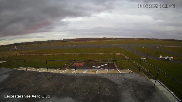 A view from a webcam at Leicestershire Aero Club, showing a wide green field under a cloudy sky. In the foreground, there is a flat rooftop with a boundary railing. Below, a runway is visible with a small aircraft taxiing on the side. Along the edges, there are grass areas and a white fence. A red flag is flying nearby, indicating the area of the airport. The lighting suggests an overcast evening.