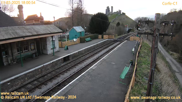 A view of a train station platform at dusk. The platform is lined with stone and has green benches. To the left, there is a building with a slate roof and a chimney. A wooden fence surrounds the area, with signs indicating "WAY OUT." In the background, there is a hill with ruins of a castle on top. Several railway tracks run alongside the platform, leading into the distance. Trees and shrubs are visible along the edges of the scene. The sky is lightening with hues of evening.