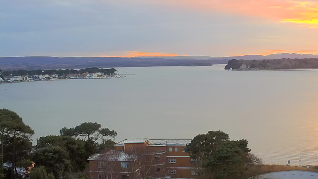 A scenic view of a calm body of water at dusk, with soft orange and pink hues in the sky as the sun sets. In the foreground, there are trees and a low building with a flat roof. To the left, a shoreline features various houses and boats, while distant hills are visible along the horizon. The overall atmosphere is tranquil and picturesque.