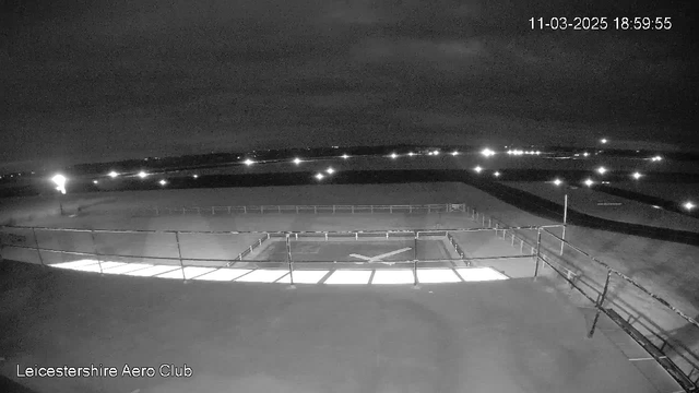 A dark, night-time scene from a webcam at the Leicestershire Aero Club. The view shows an airfield with a illuminated helipad marked by white lines and a dark background. There are faint lights in the distance, possibly from buildings or vehicles. The sky appears cloudy, creating a moody atmosphere with limited visibility. The timestamp in the corner indicates the date and time as March 11, 2025, at 18:59:55.