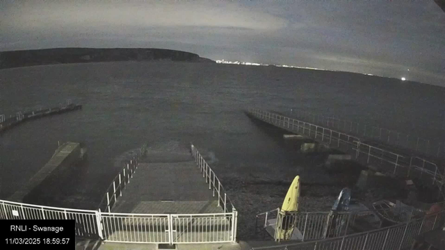 A view of a dark shoreline with water reflecting light. A long walkway extends from the bottom of the image towards a pier on the left side. There are two kayaks visible on the right side of the image, with one yellow and one blue. The sky is overcast with some low clouds, and lights from a distant area can be seen glinting on the horizon. The overall scene suggests evening or twilight conditions.