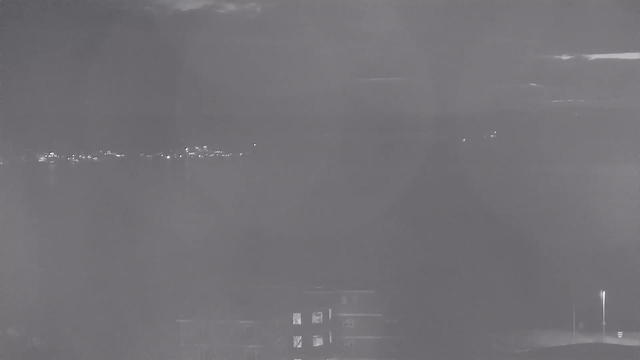 A night view from a webcam showing a dark, cloudy sky above a body of water. In the distance, faint lights from buildings and possibly a bridge can be seen along the shoreline. The foreground is mostly dark, with some outlines of structures, including a building with illuminated windows. There is also a faint light from a streetlamp to the right, casting a soft glow. Overall, the image conveys a serene, nighttime atmosphere.