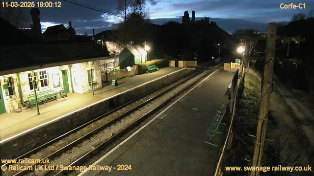 A dimly lit railway station platform at dusk, with a stone building featuring large windows and a green bench. The platform is lined with gravel and has wooden fences on either side. In the background, there are silhouettes of trees and a hill with structures. A street lamp casts a warm glow, while a sign indicating "WAY OUT" is visible. The tracks are empty, leading away from the station.