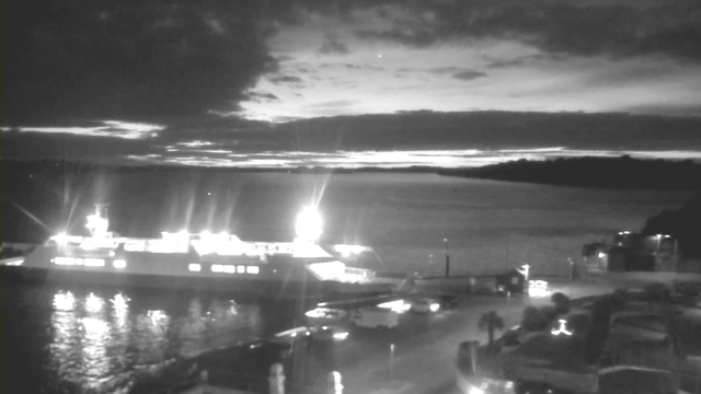 A dark, nighttime scene featuring a large ferry docked near the waterfront. The ferry is illuminated with bright lights, reflecting on the water's surface. The background includes a cloudy sky with various shades of gray, and the horizon is barely visible. Some lights from buildings and vehicles are seen along the shore, and a boat is positioned nearby.