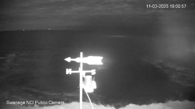 A black and white webcam image showing a view of the ocean with dark, cloudy skies. A directional weather vane is visible on the left side, indicating north and south. The ocean meets the horizon, with faint outlines of waves. The date and time displayed at the top right reads "11-03-2025 19:00:57." The bottom left corner signifies "Swanage NCI Public Camera."