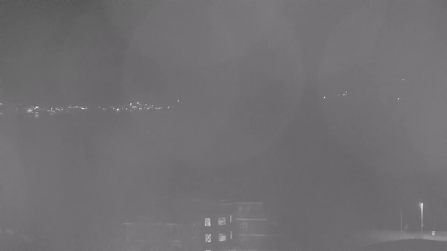 A grayscale image captured by a webcam shows a hazy landscape at night. In the distance, there are faint lights visible, possibly from buildings or street lamps. The foreground includes a partially visible building with lit windows, while the overall scene appears foggy or unclear, suggesting a low visibility condition.