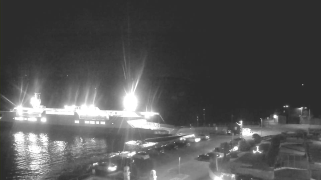 A dimly lit scene featuring a large ferry docked at a harbor, illuminated by bright lights. The water reflects some of the light from the ferry. There are several parked cars along the waterfront and a pathway leading towards the ferry. The background is dark, indicating it is nighttime, with a few distant lights visible.